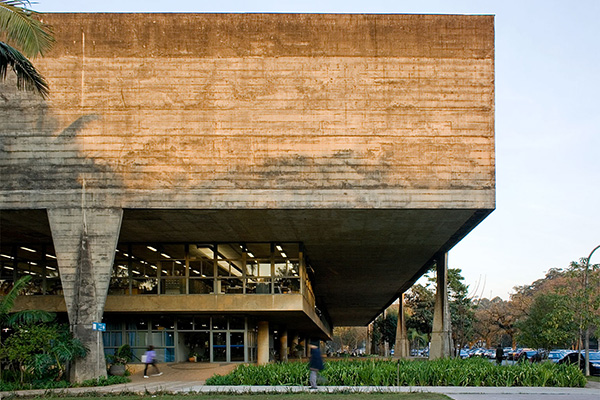front facade of a building built in exposed concrete, frontal light at the end of the day,
     triangular pillars. classic architecture photo with some people walking in front of the building but out of focus due to the movement. faculty of architecture and urbanism at usp,
      designed by Vilanova Artigas.