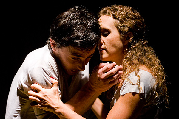 an actor and an actress, in a contrasting and dramatic upper 
    light, in front of a black background, very close to each other. She holds him tightly by the arms while he looks a little scared. Eduardo okamoto and Alice possani are acting in the play Chuva Pasmada
    by Mia Couto.